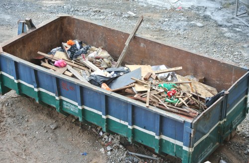 A large, rectangular metal skip bin placed on a gravel surface, used for outdoor waste removal in a garden clearance context. The container has a rust-colored interior and is filled with various construction debris, including wooden planks, broken pieces of plywood, plastic bags, and small miscellaneous rubbish items. Some materials are leaning against the sides of the skip, with the wooden planks scattered unevenly, showing a range of textures from smooth painted surfaces to rough, splintered edges. Surrounding the skip is a gravel driveway or yard area, with loose stones and dirt visible on the ground, suggesting an active site for rubbish collection and disposal services, such as those offered by Garden Clearance Addiscombe near Croydon. The overall scene reflects a typical outdoor waste removal setup, emphasizing the importance of proper rubbish management on residential or commercial premises in the local area.