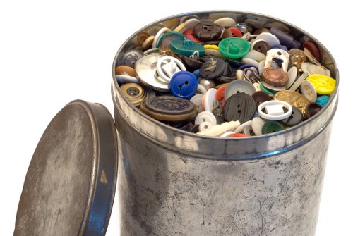 A cylindrical metal container with a weathered, slightly dented surface, filled to the top with a collection of assorted plastic and metal buttons in various sizes, shapes, and colors, including blue, white, black, beige, and brown. The container is positioned on a plain white background, with a round, metallic lid placed beside it, tilted slightly to reveal the circular inner surface. The image captures the textures of the metal, the glossy and matte finishes of the buttons, and the diversity of colors, suggesting a focus on waste or rubbish collection, which may relate to rubbish removal services offered by Garden Clearance Addiscombe, situated in the postcode area near London. The lighting is even, with soft shadows indicating a neutral environment, emphasizing the collection of buttons as the central visual element relevant to waste disposal or recycling themes.