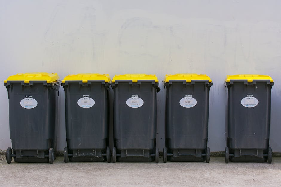 A set of three large wheeled rubbish containers positioned against a brick wall on a city street. The two red bins, labeled with a 'Recyclables' sign, are designated for recyclable waste and stand on 