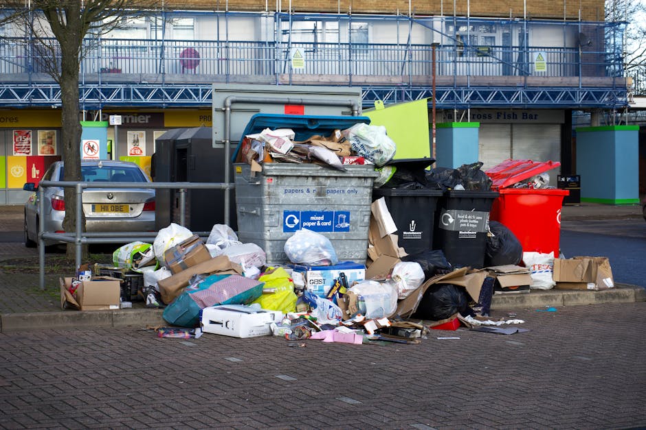 An aerial view of a large outdoor rubbish accumulation site showing three individuals sorting through piled waste. The scene features an area densely filled with various types of discarded items inclu