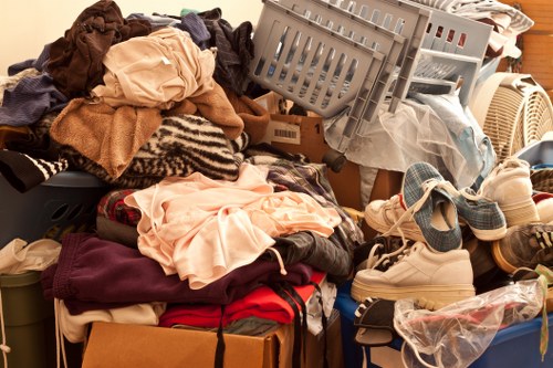 A large collection of mixed household items and clothing, including folded and unfolded garments in various colours such as peach, red, beige, and dark tones, piled on top of each other on a surface indoors. Among the clothing are shoes, with a pair of light-colored sneakers visible in the foreground, and plastic bags containing additional items. Overlapping boxes and plastic laundry baskets, some filled with textiles, are positioned behind the pile, with one basket tilted at an angle. The background features a wooden surface, possibly a furniture piece or part of a room interior, and the lighting appears natural or soft, emphasizing the disorganized state typical of items to be collected for rubbish removal or clearance in an Addiscombe property. Garden Clearance Addiscombe would handle such clutter efficiently, facilitating clean and organized rubbish disposal in the local area.