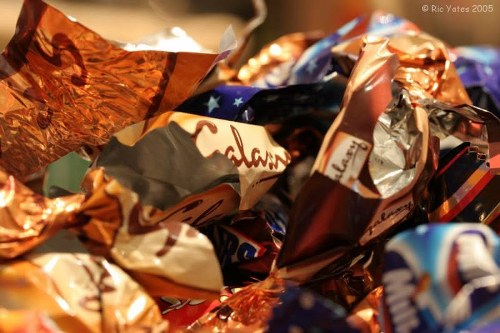 A close-up image of a pile of crumpled and twisted foil wrappers and plastic packaging, featuring metallic gold, silver, and various colored sections, likely from confectionery or snack products. The wrappers are piled together in an unorganized manner, with some partially opened or torn, revealing creased and reflective surfaces that catch light in different directions. The background is blurred, suggesting they are gathered in a container or on a surface, possibly for rubbish collection or disposal. The scene is well-lit, emphasizing the shiny textures and vibrant colors of the wrappers, which are typical items handled during a rubbish removal service in Addiscombe. This imagery aligns with the waste management theme of disposing of packaging waste, as handled by Garden Clearance Addiscombe, in a professional and factual manner reflective of local rubbish clearance tasks.
