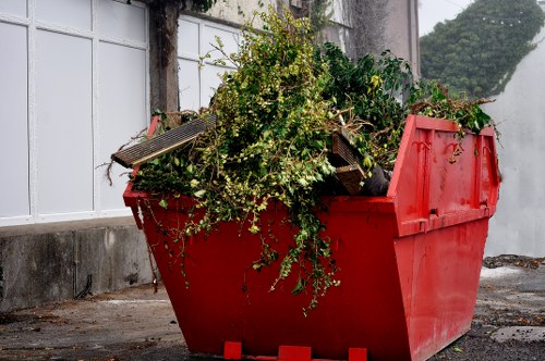 A large, red plastic skip with high sides is positioned on a paved outdoor area near a concrete wall and white-framed glass windows. The skip is filled with green garden debris, including leafy branches and small shrubs, with some broken wooden planks and pieces of fencing timber visible among the foliage. The surface of the debris appears fresh and unwound, indicating recent clearing activity. The background features a concrete wall with a white window frame and a blurred view of trees or foliage further behind, suggesting the scene takes place in a residential or garden setting. The lighting is natural, with diffuse daylight illuminating the scene, emphasizing the vibrant red of the skip and the fresh green of the garden waste. This image is relevant for a rubbish removal service such as Garden Clearance Addiscombe, highlighting their capacity to handle garden waste and debris cleanup in local properties within the Croydon or Addiscombe area.
