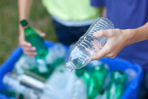 A close-up view of a person's hand holding a clear plastic PET bottle over a blue recycling bin filled with various other plastic bottles and recycling materials. In the background, another individual is visible, wearing a yellow and blue top, standing outdoors on a paved or grassy area. The scene depicts a waste collection or recycling activity, emphasizing the handling and sorting of recyclable plastics, relevant to rubbish removal services in areas such as Addiscombe. The plastic bottles are transparent or green, with some showing ridges or textures typical of soft drink containers, and the environment appears well-lit with natural daylight. This image illustrates environmentally focused rubbish disposal practices, as managed by local waste management services like Garden Clearance Addiscombe.